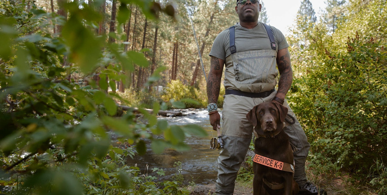 Chad in fly fishing gear and dog axe in service dog vest pose by the river.