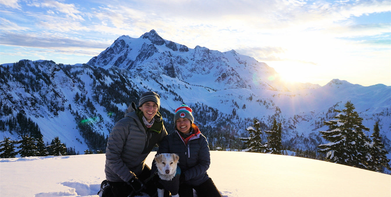 A woman and man sit with their dog on a snowy mountain.